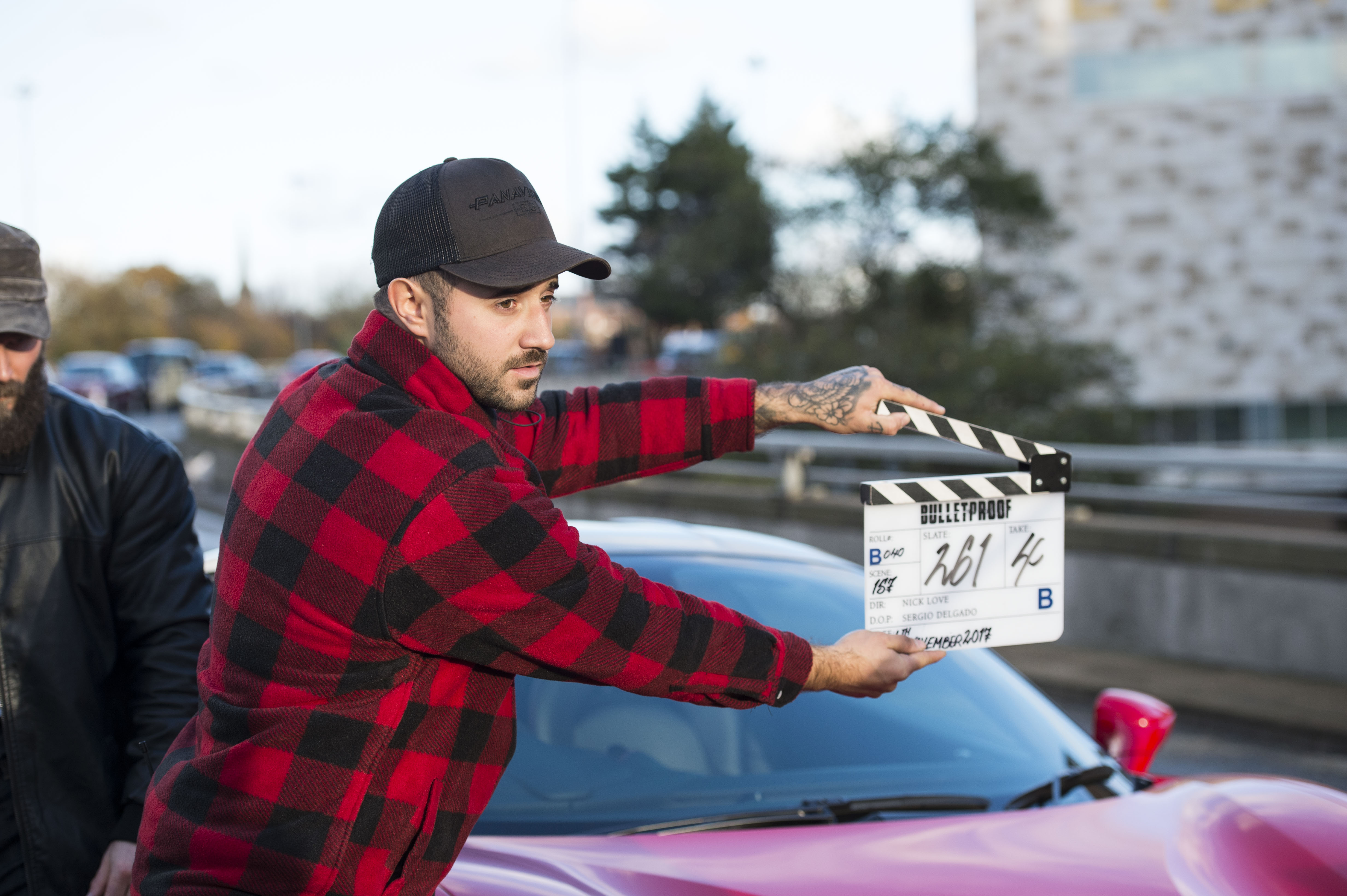 A man wearing a red checked shirt and baseball cap holds a clapper board in front of a sports car