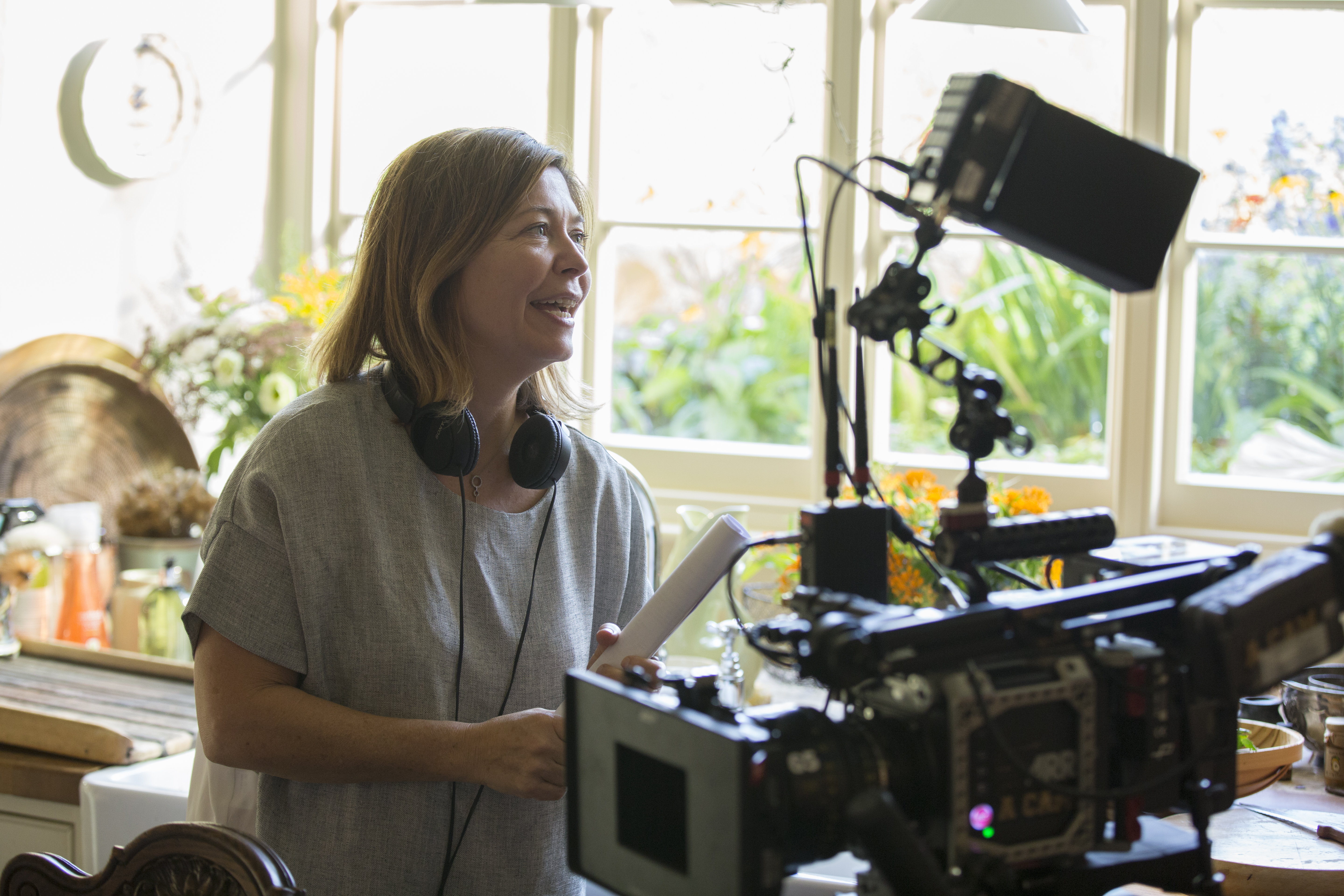 A woman wearing headphones stands behind a camera giving instructions