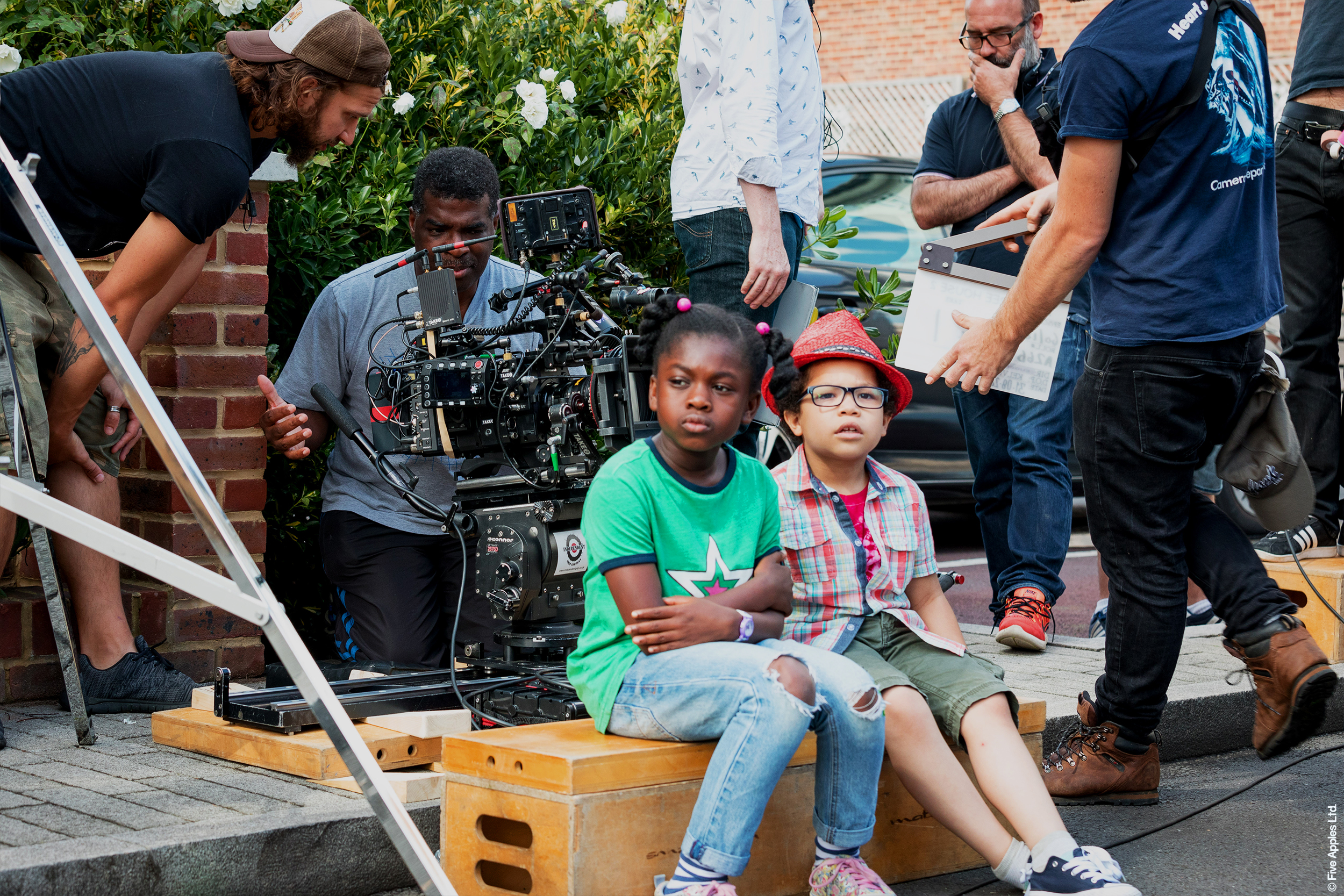 Two children sit on a wooded box surrounded by production crew on the set of Apple Tree House