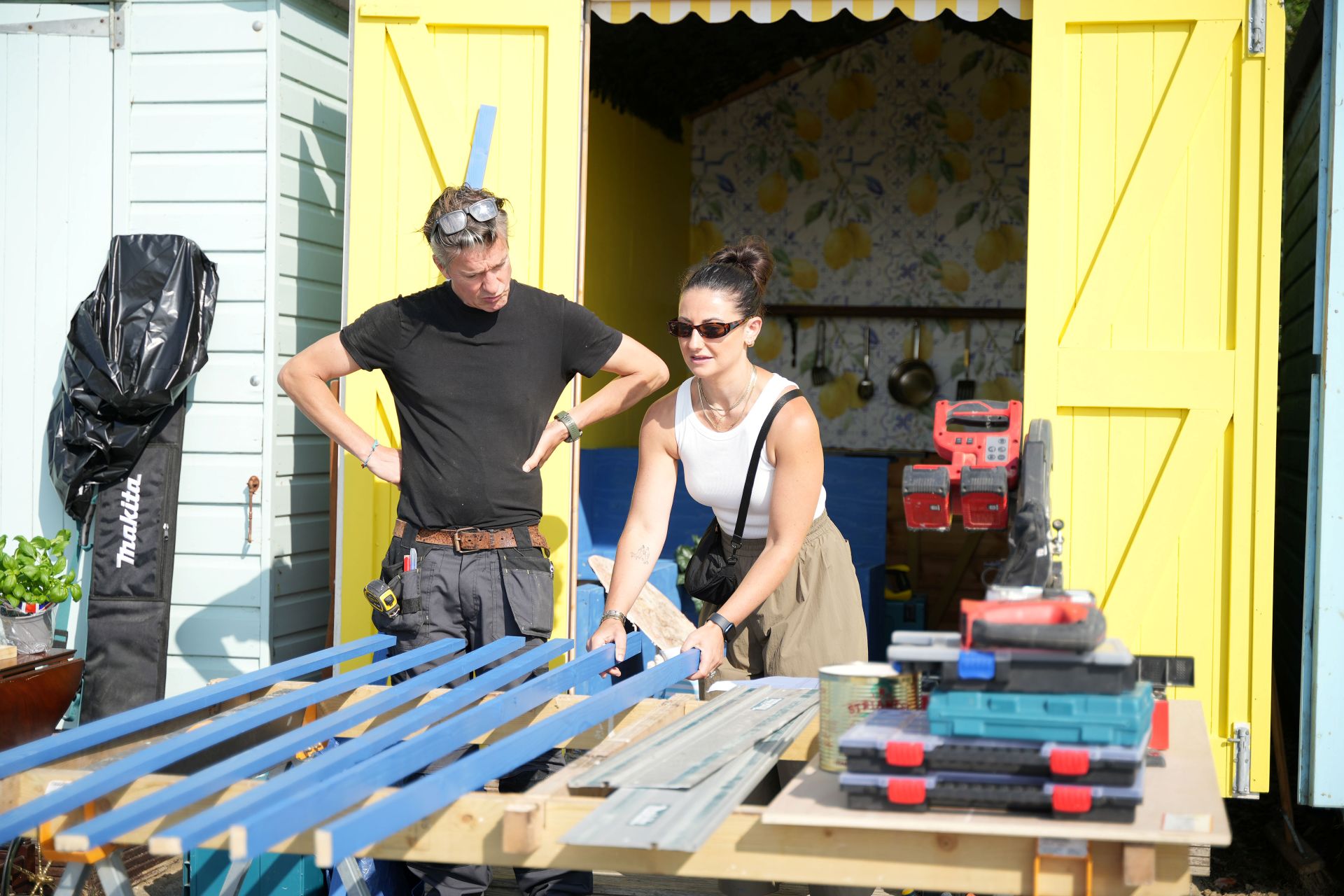 Two people stand in an outdoor workshop, working on large wooden panels