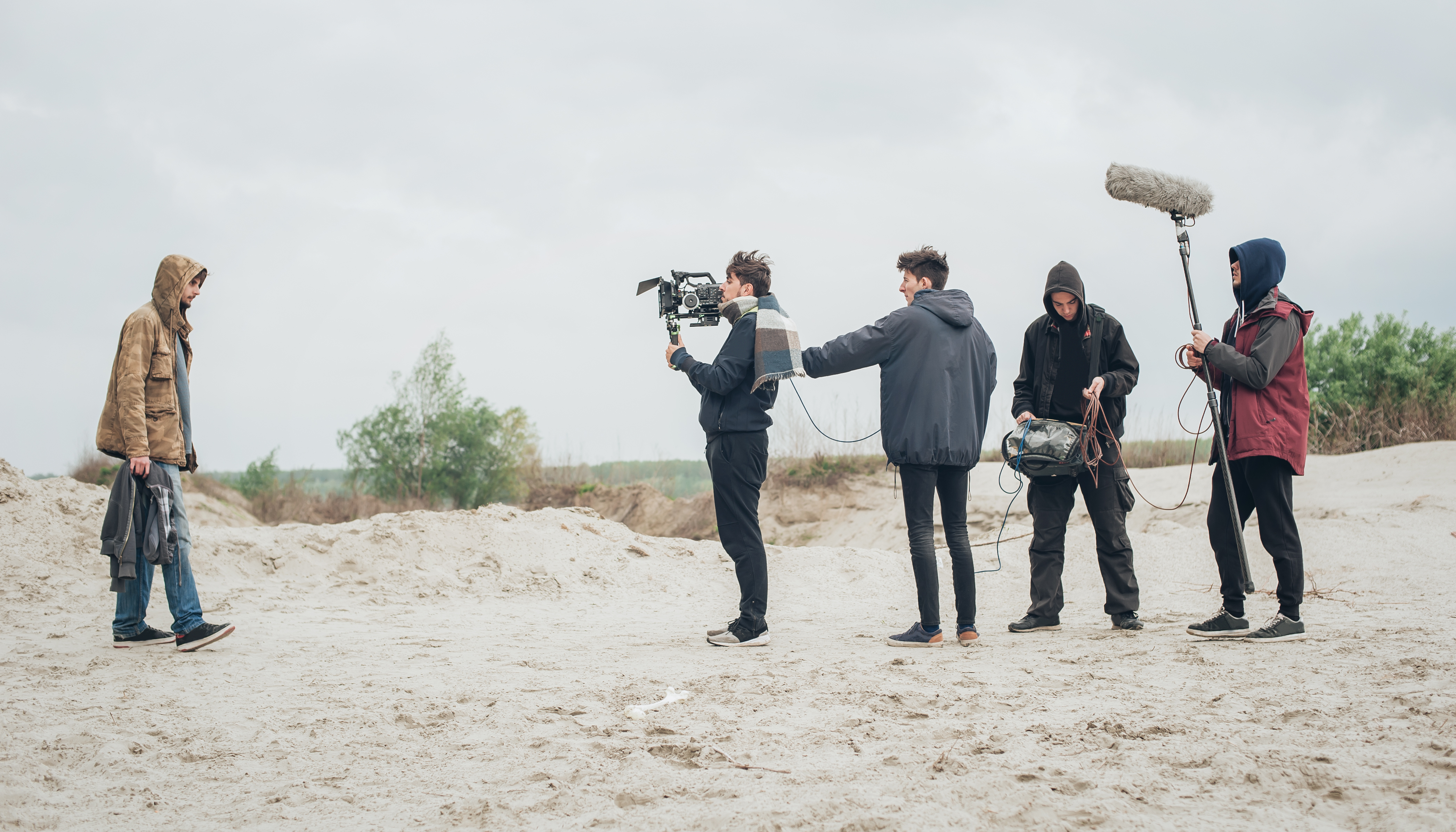 Camera and sound crew film a man in a hooded coat and trainers walking on a beach