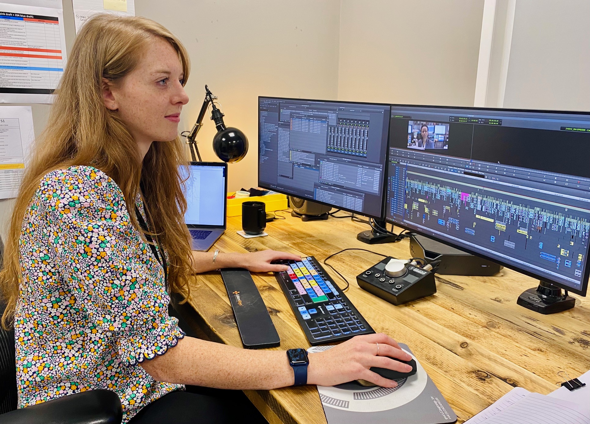 A woman sits at a desk in front of a set of computer monitors editing a sequence in a video editing software.