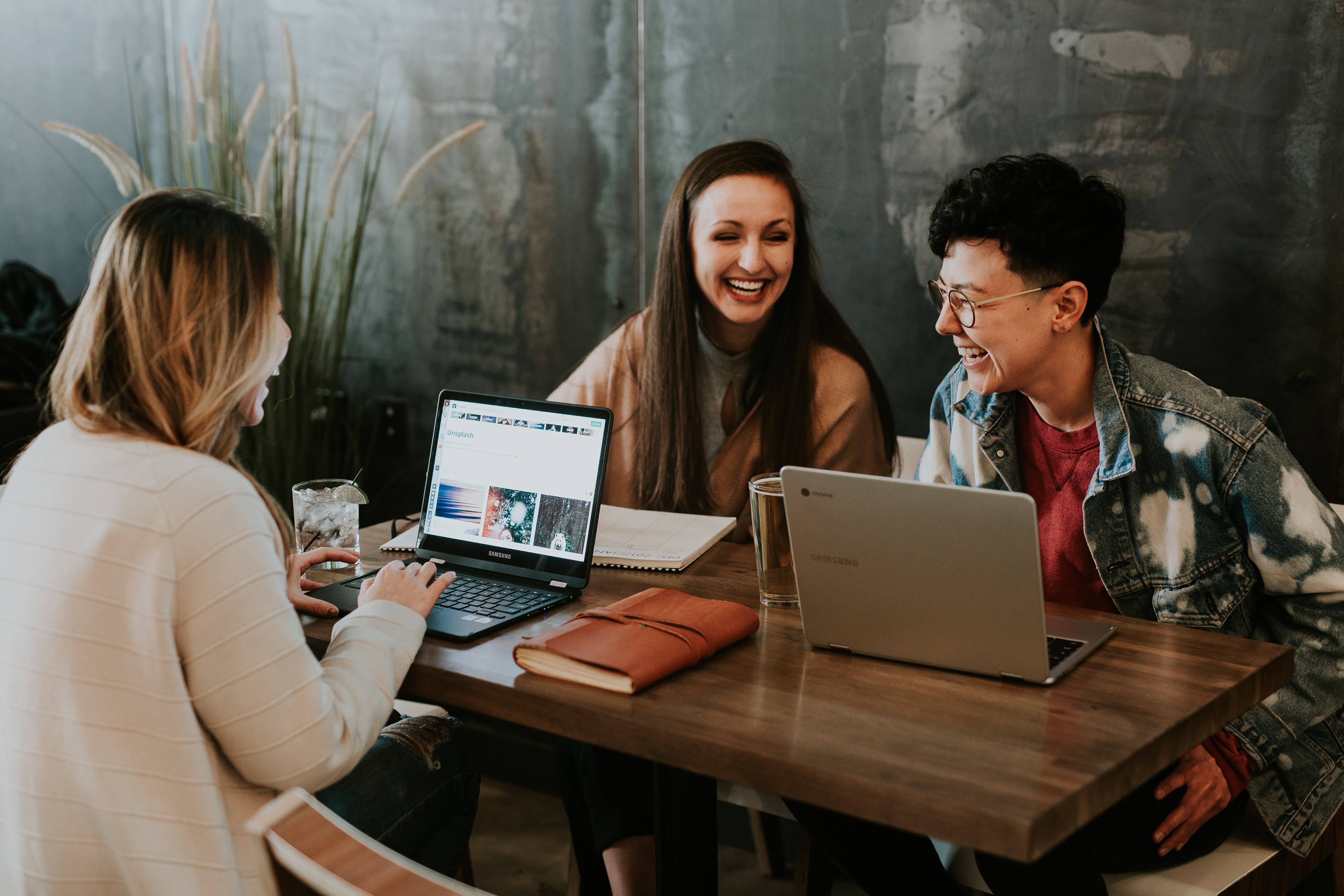 Three people sit around a table with laptops, laughing together