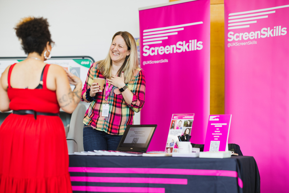 Kate Efomi stands behind a desk in front of a ScreenSkills logo banner talking to someone at the BAFTA Guru live event in Scotland