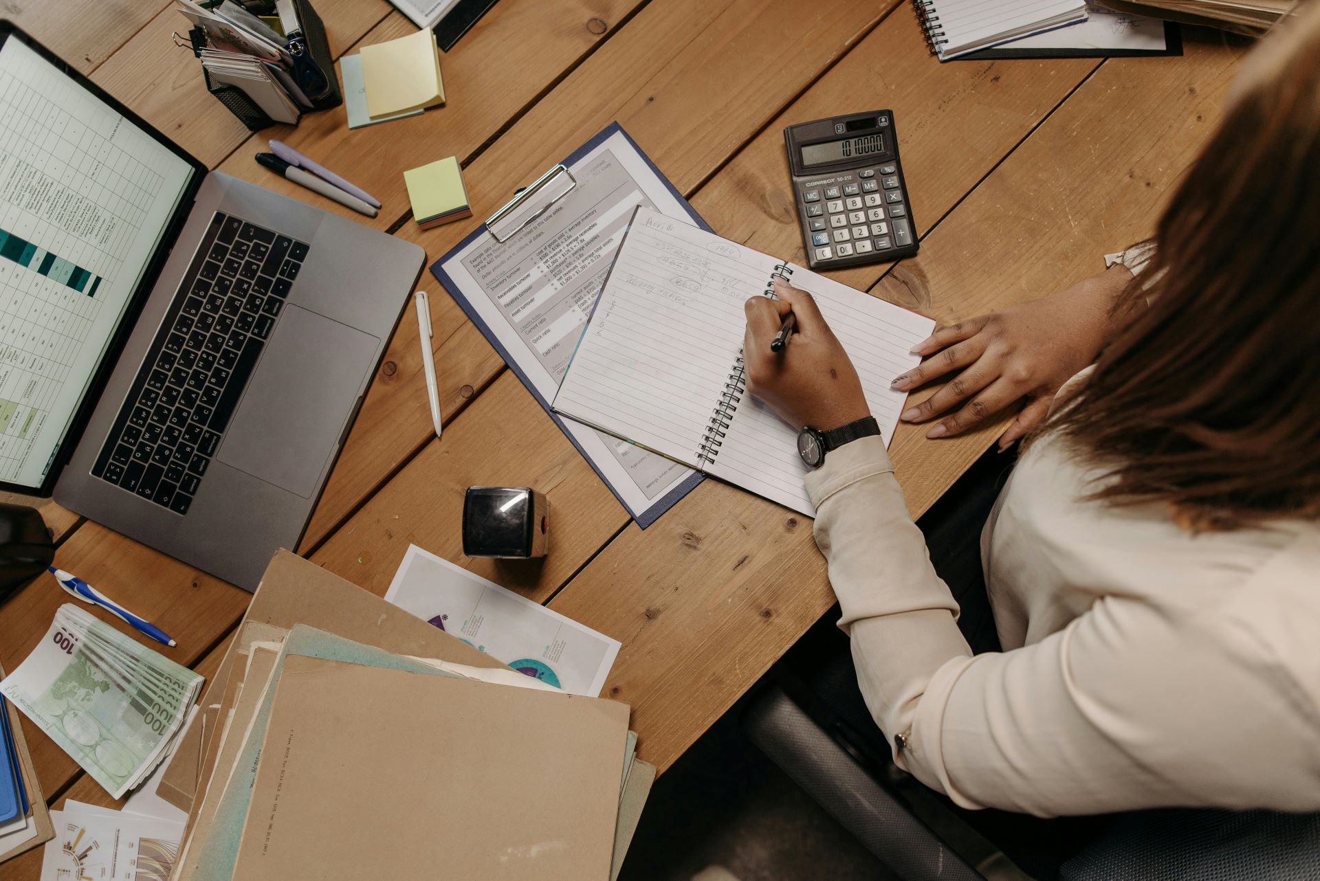 Woman sitting at a desk with an open laptop, folders and a calculator, writing into a notebook