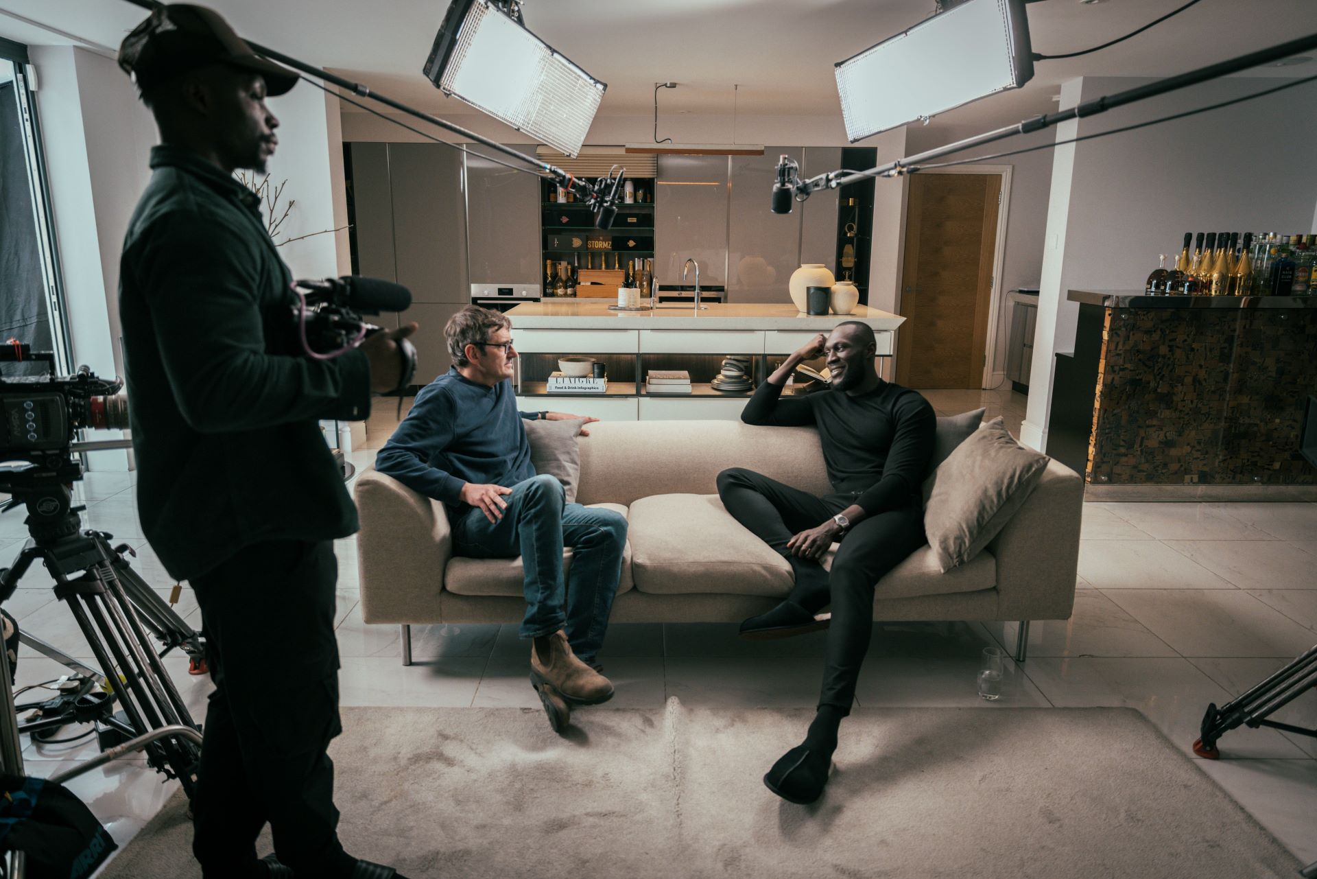 Louis Theroux and Stormzy sit chatting on a sofa surrounded by filming equipment. A cameraman holding a camera stands off to one side.