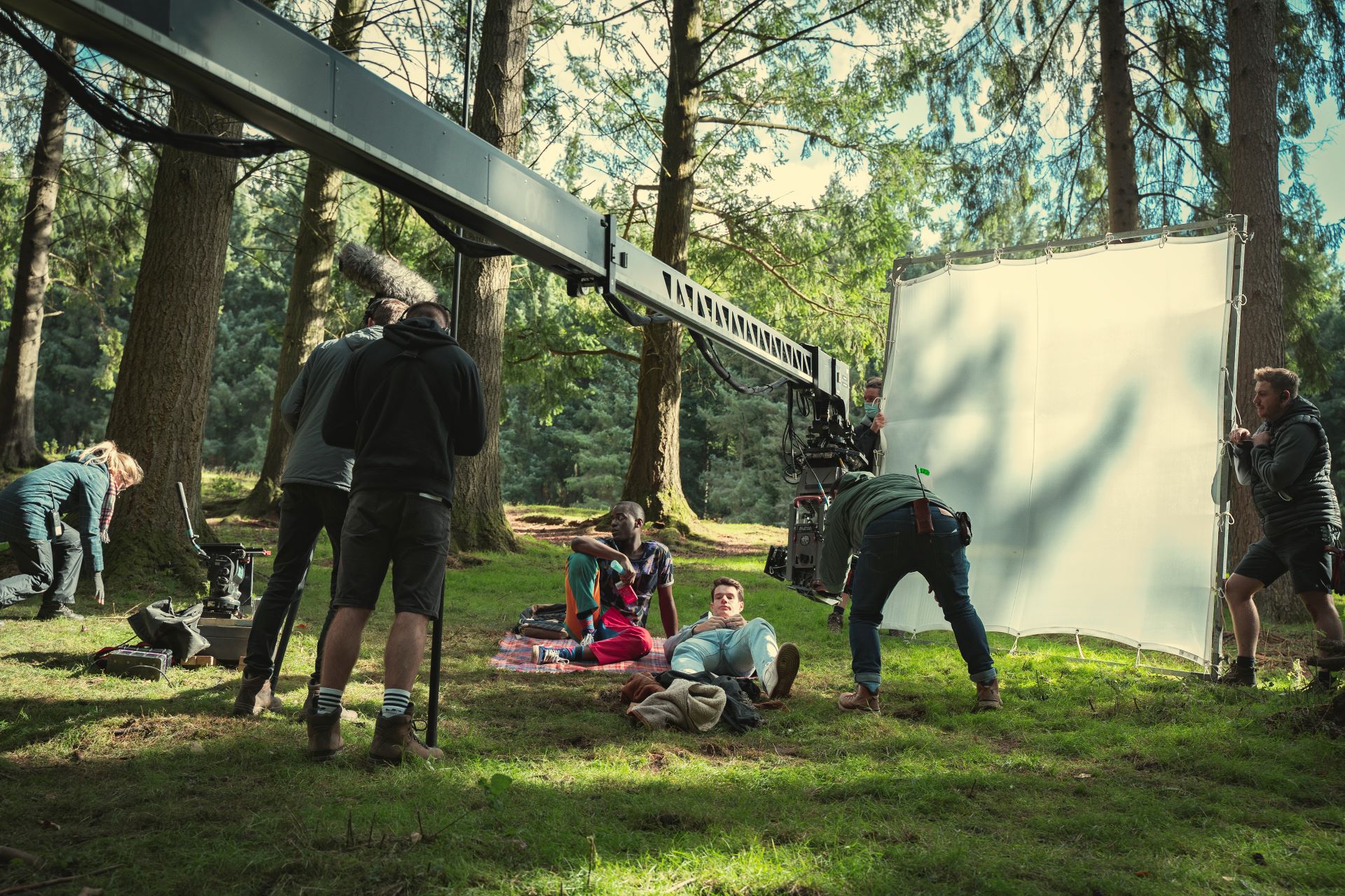 On location for Sex Education season 3, two actors lounge on picnic blankets in a forest in front of a large white screen and a camera boom 