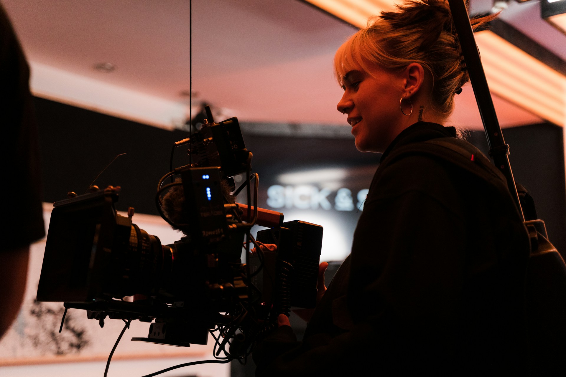 A woman holds a large camera which is strapped to her on a film set