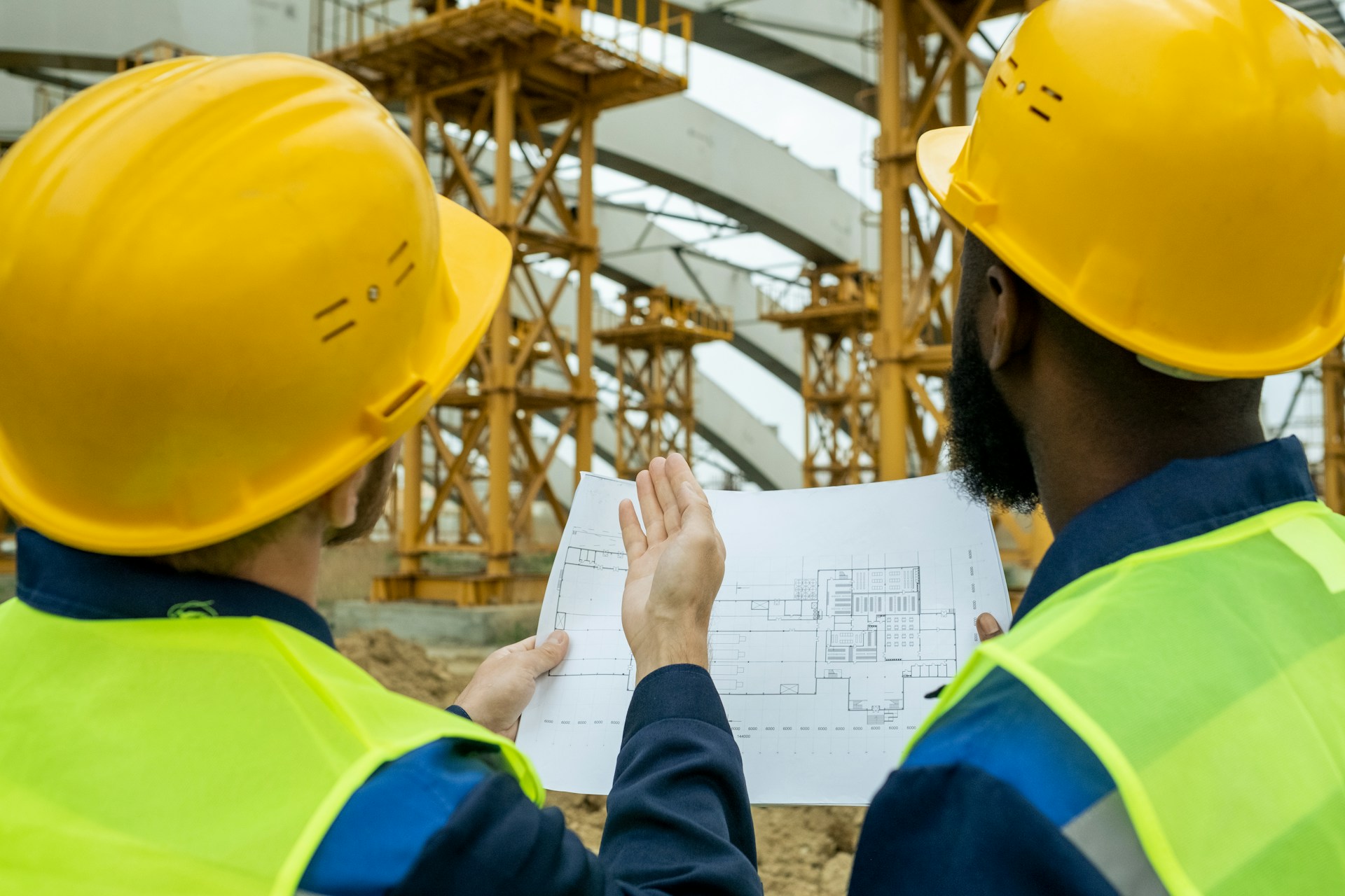 Rear view of two workers standing outdoors in work helmets comparing the blueprint with the building under construction