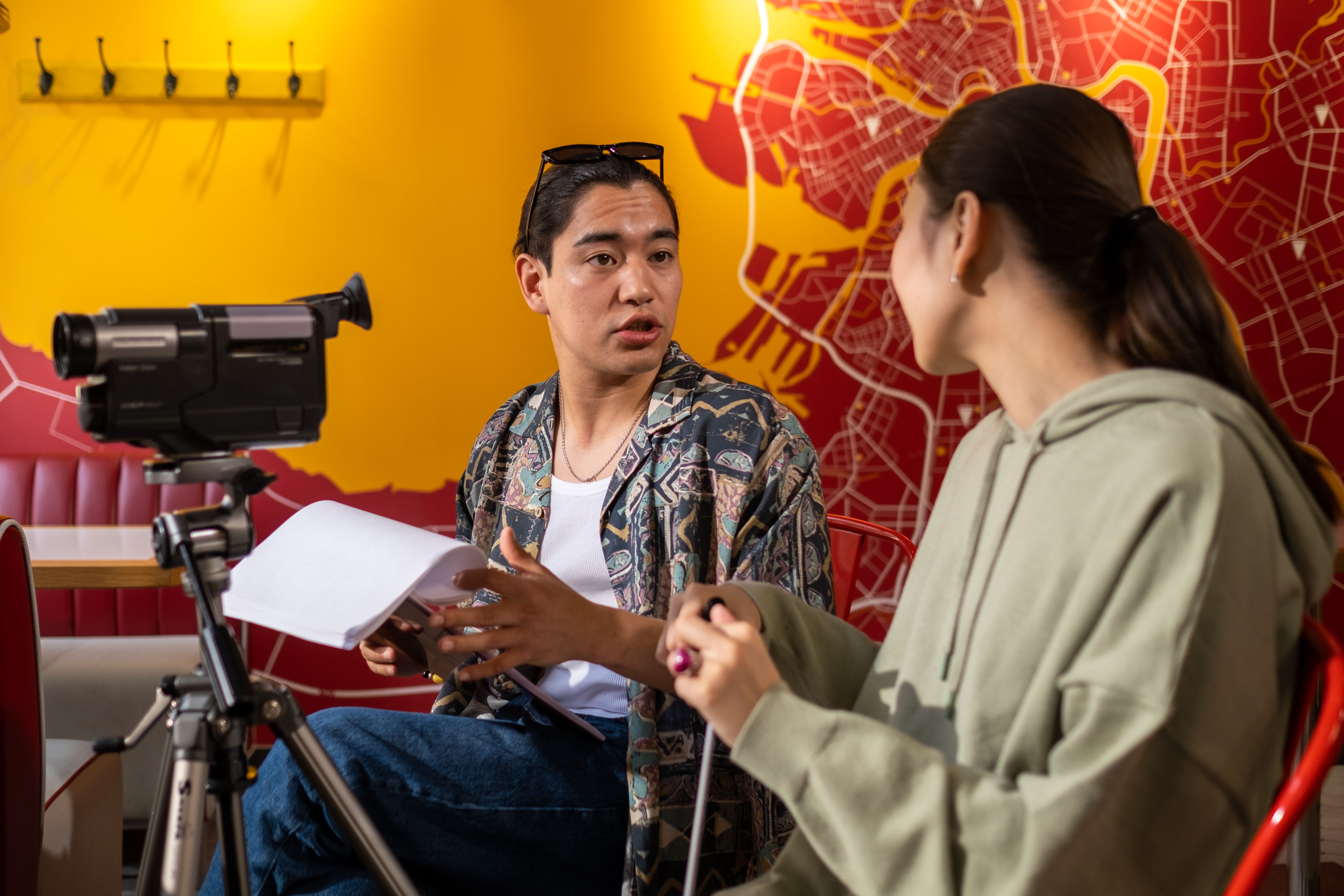 Two people sit discussing a script. To the left of them is a camcorder mounted on a tripod