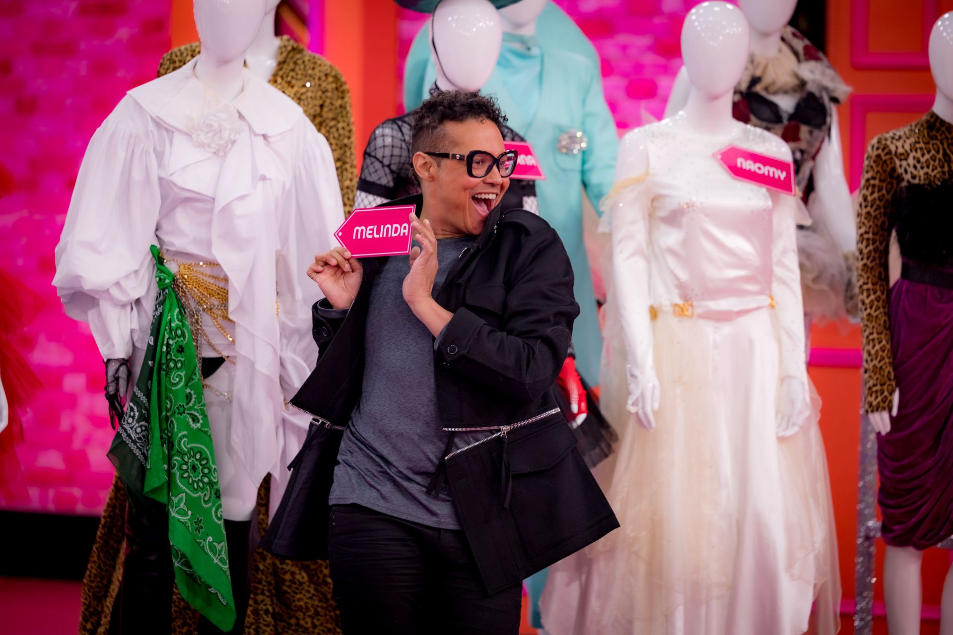 Someone posing holding a name card. Rows of mannequins wearing dresses stand in the background.