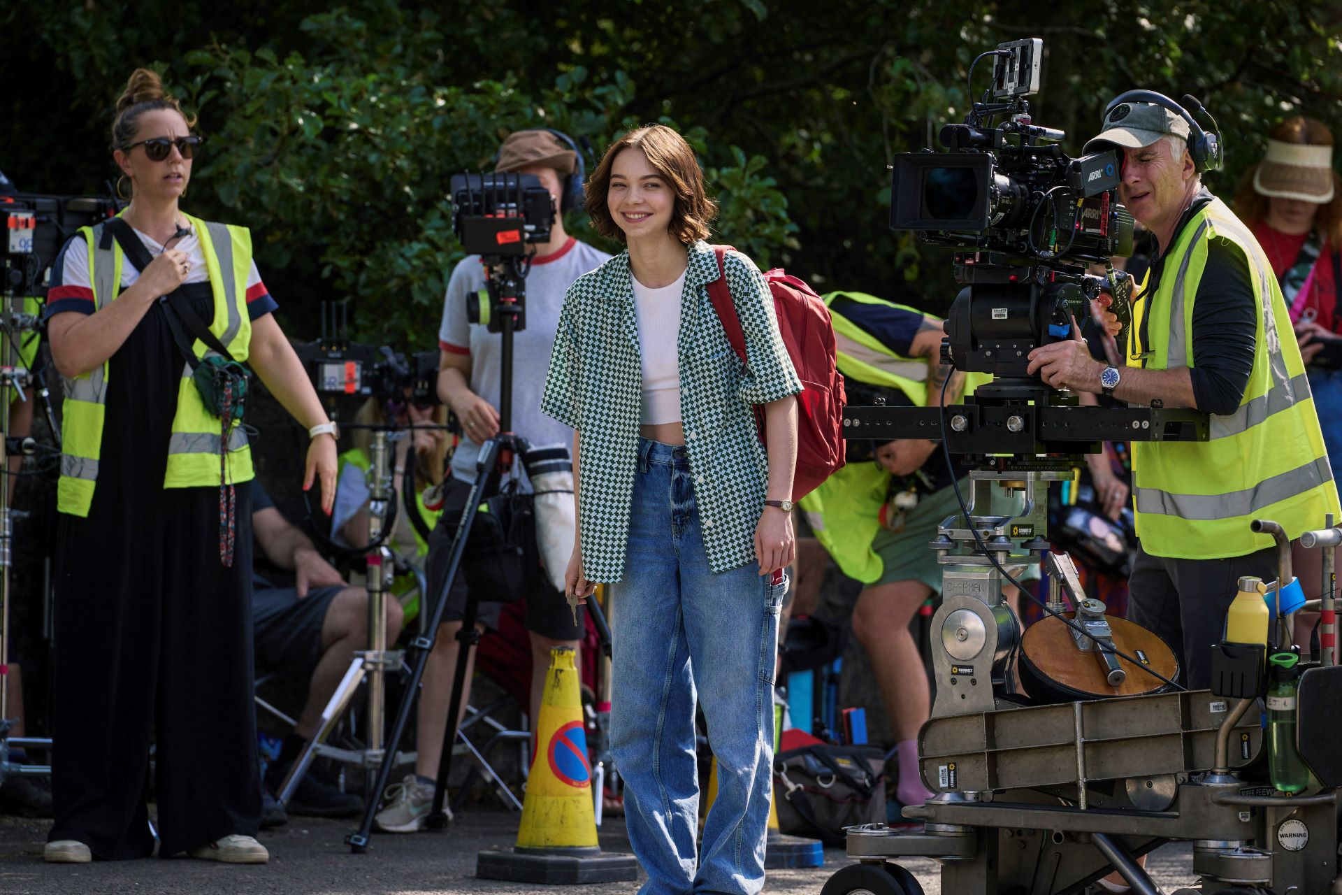 Emma Myers stands in the centre of an outside set on A Good Girl's Guide to Murder production. Surrounding her is camera equipment being operated by various production crew members in high-vis jackets.