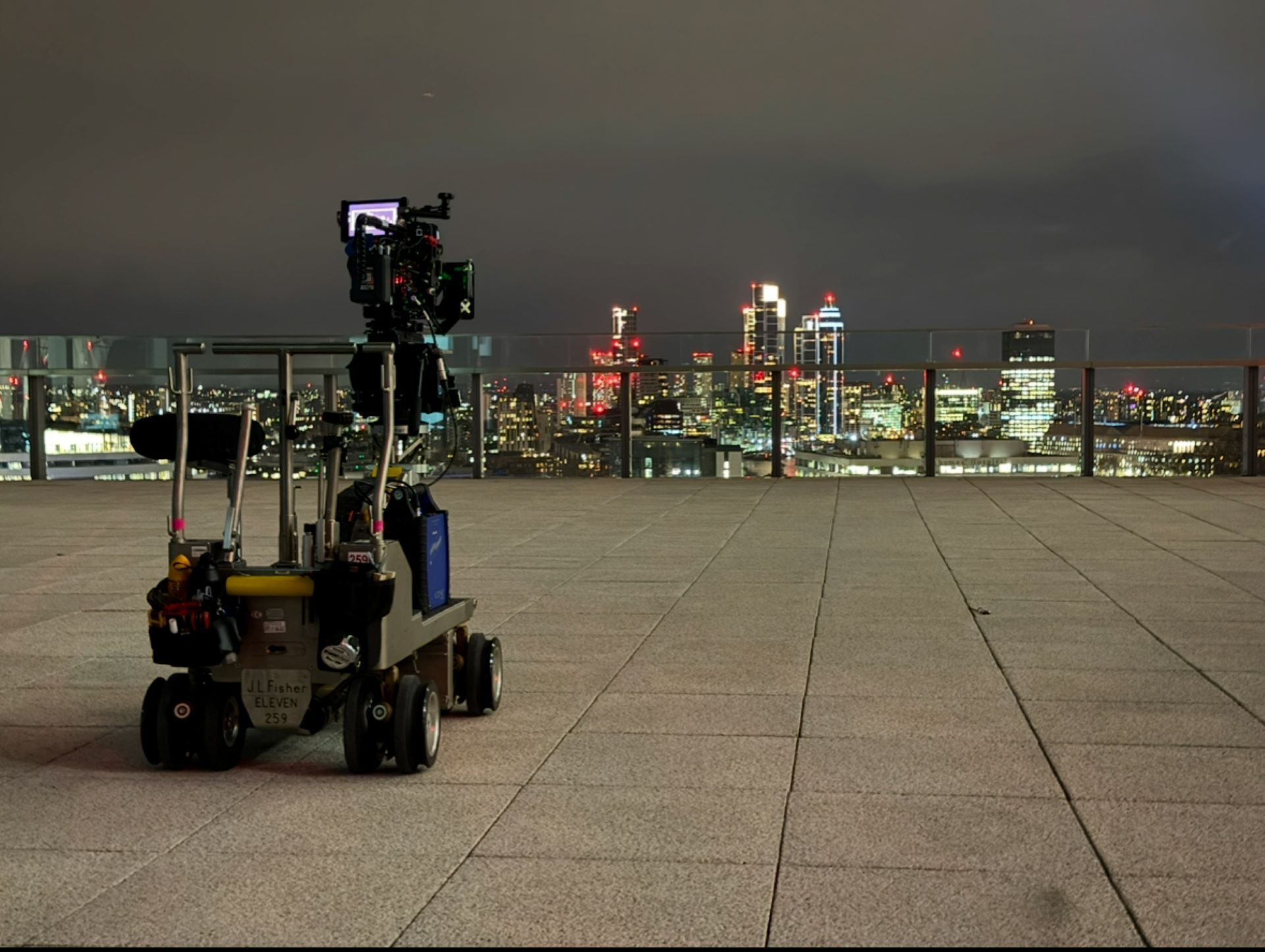 A camera stands on a moving platform on a roof overlooking a nighttime cityscape