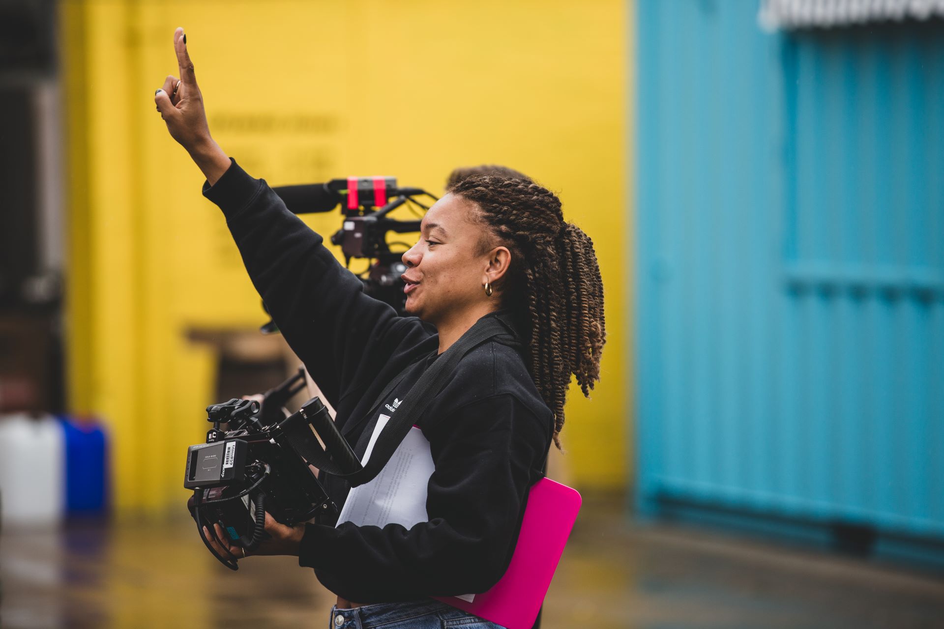 A woman stands on location holding a camera with her arm in the air