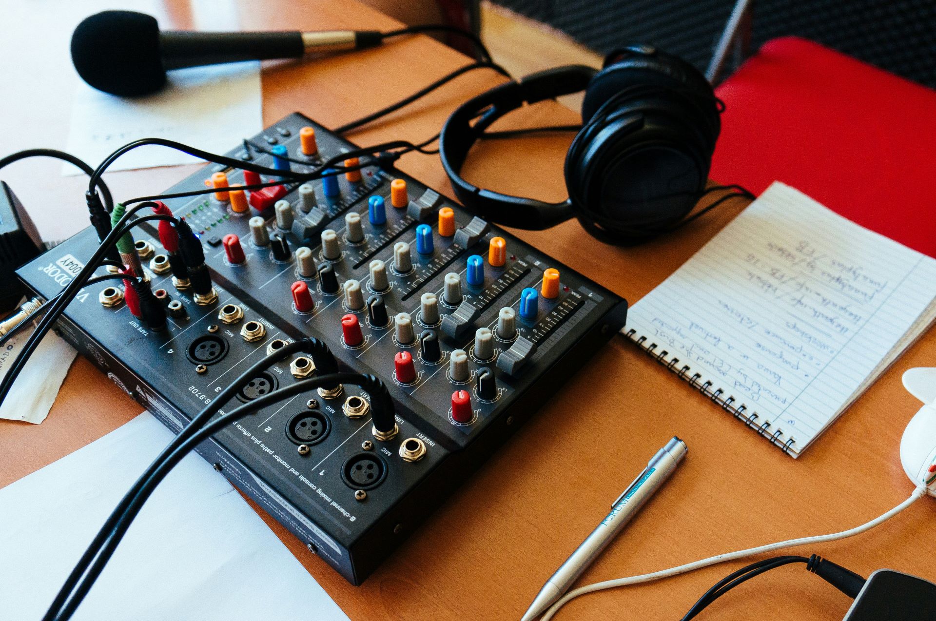 A picture of an audio mixer on a desk. Also on the desk is a notebook and pen, a microphone and a pair of headphones.